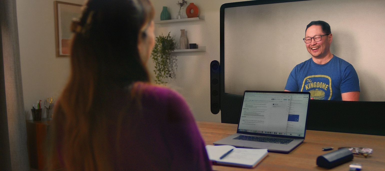 Two people in conversation using Beam. Person smiling on Beam display while engaged in conversation. The back of the head and hand of person in the foreground. There is a laptop and eyeglasses on the table between them.