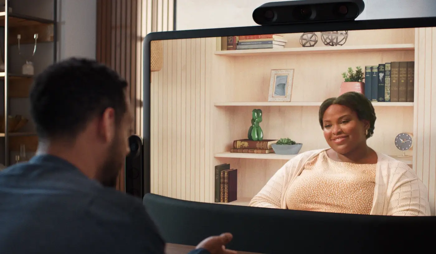 Person seated at laptop in office stands up and checks smartphone screen. Person seated on bench with headphones on looks at laptop screen with hands clasped. Person seated at desk alone in office types on laptop. Two people talk and smile at conference table with laptops open. Two people using American sign language have a conversation seated on the same side of an office table. One person signs “That’s not a good idea”. Close up of person’s face while signing. Person smiling in response. Close up on seated person talking to another person in a seat. They gesture in conversation. Camera moves back to reveal the Beam display and table and that they are using Beam for their conversation. We see both individuals on the Beam display gesturing and smiling in conversation.
