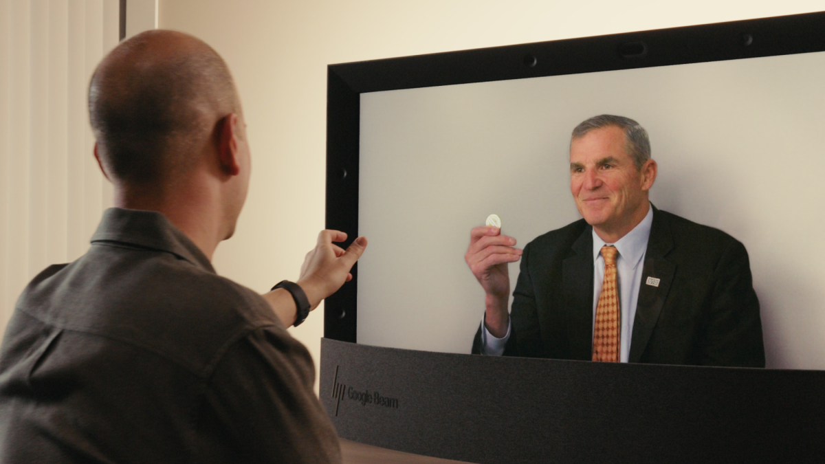 A man, viewed from the back, reaches toward a Google Beam display. On the screen, a man in a suit and tie smiles and holds up a small, round, silver token.