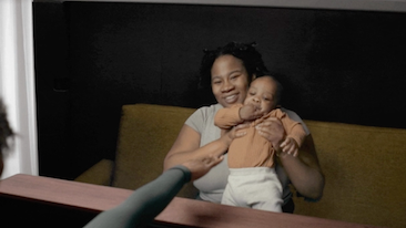 Two people in conversation on Project Starline, one is smiling and holding up a baby on Starline display. The other person reaches towards the pair.