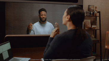 Two people in conversation using Starline, the person on Starline display holds up their hands up expressively. The back of the head can be seen of the person in their office.